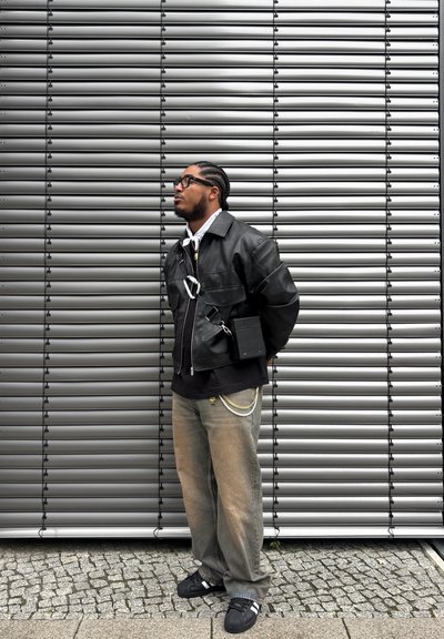 Chaqueta de cuero negra con un broche plateado, superpuesta a una camiseta negra. Jeans holgados de color gris claro con cadenas como accesorios, zapatos negros, contra un fondo gris.