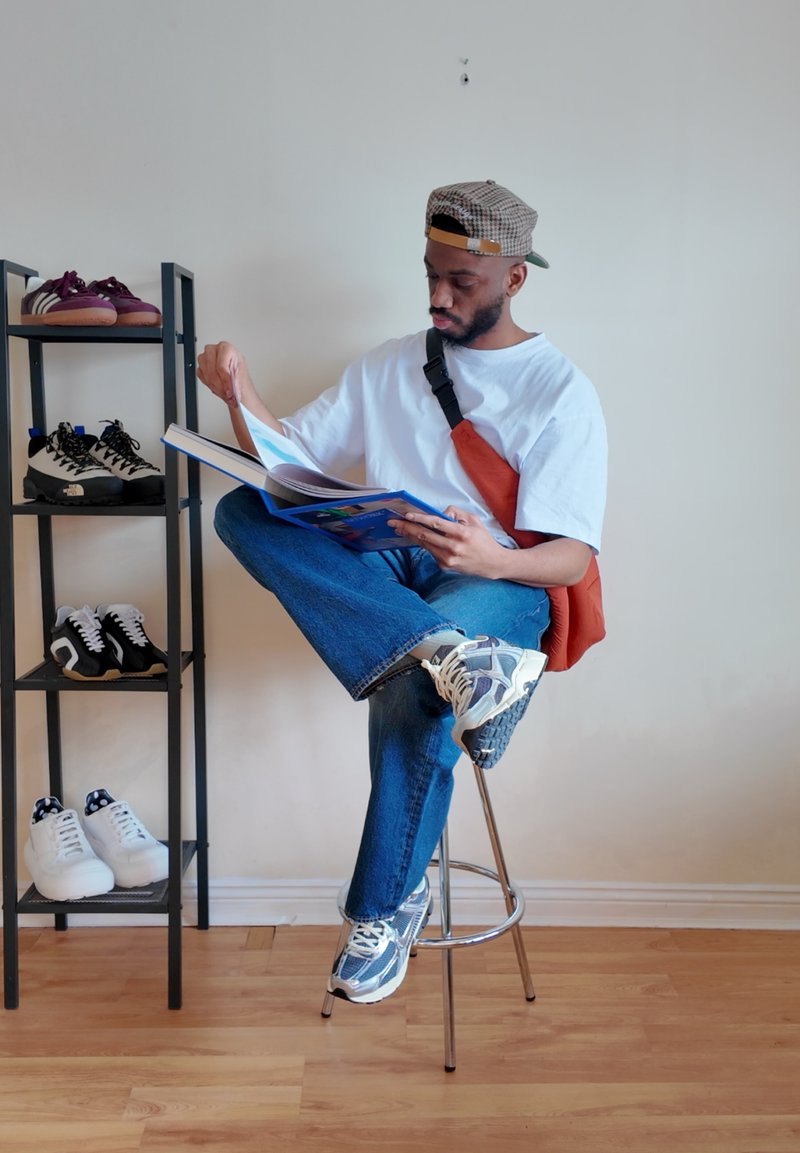 Homme portant une casquette et un sac en bandoulière assis sur un tabouret, lisant un livre à côté d'une étagère contenant quatre paires de chaussures.