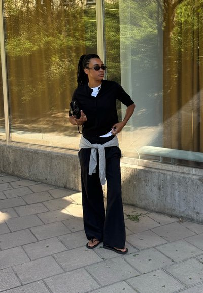 Camisa negra de botones, camiseta blanca, suéter gris atado a la cintura, pantalones holgados negros, gafas de sol negras, sosteniendo una pequeña bolsa negra.