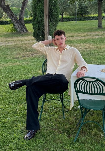 Camisa de lino beige claro con las mangas arremangadas, pantalones negros y zapatos negros brillantes. Sentado en una silla verde, con una postura relajada.