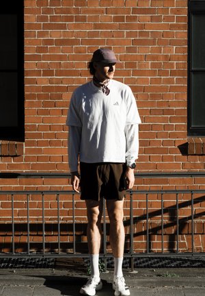 Hombre con camisa blanca de manga larga, pantalones cortos oscuros, gorra y zapatillas de deporte, de pie junto a una pared de ladrillo iluminada por el sol con una barandilla de metal delante.