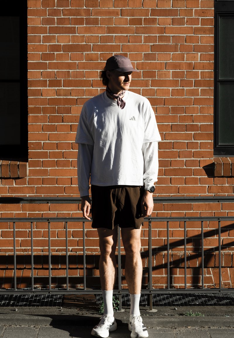 Mand iført hvid langærmet skjorte, mørke shorts, kasket og sneakers, stående op ad en solbelyst murstensvæg med metalgelænder foran.