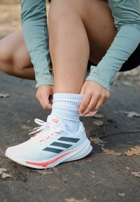 Tênis desportivos brancos com riscas pretas, detalhes vermelhos e um design em rede. Uma pessoa ajusta meias brancas enquanto se agacha num caminho.
