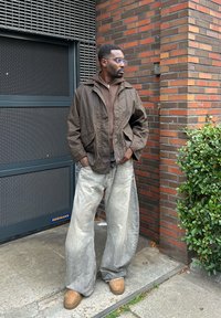 Braune Kapuzenjacke, braunes Lagenoberteil, weit geschnittene, verblasste Denim-Jeans, hellbraune Schuhe, Brille, steht gegen eine Ziegelwand.