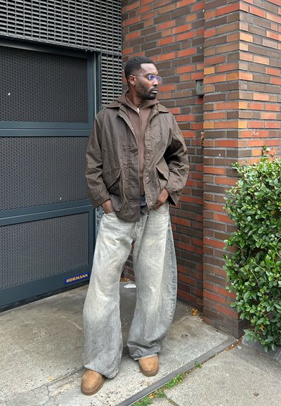 Chaqueta con capucha marrón, camiseta marrón de capas, jeans de mezclilla desgastados y holgados, calzado marrón claro, gafas, de pie frente a una pared de ladrillo.
