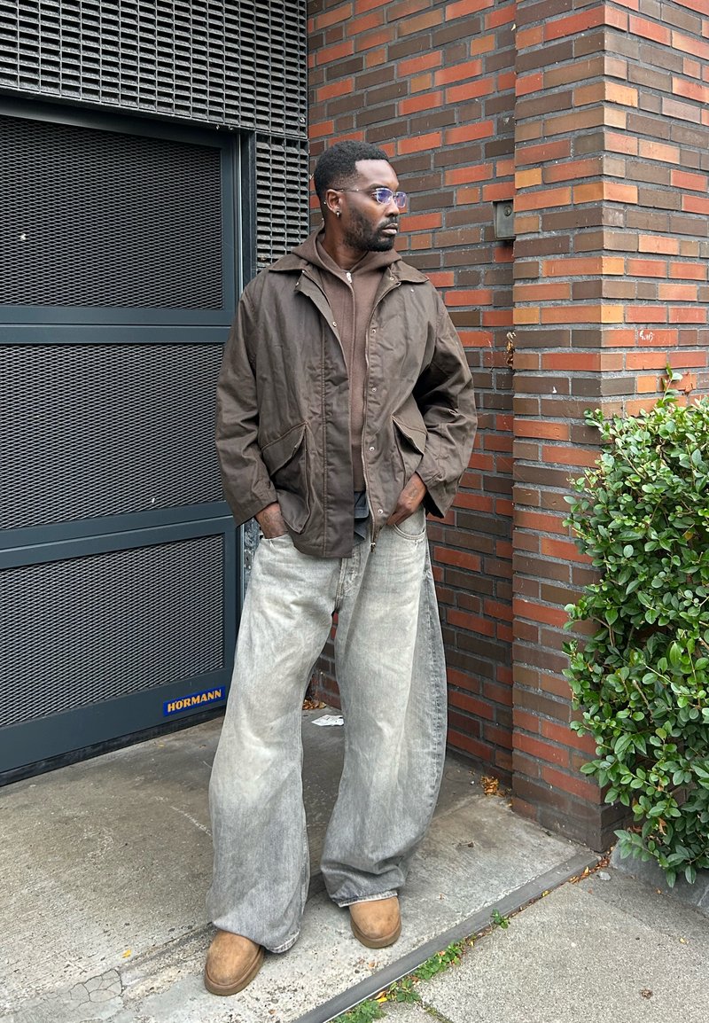 Giacca cappuccio marrone, top stratificato marrone, jeans in denim baggy sbiaditi, calzature marrone chiaro, occhiali, in piedi contro un muro di mattoni.