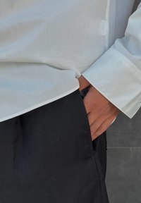 Biała koszula z kołnierzykiem o gładkiej teksturze, połączona z czarnymi, prążkowanymi spodniami. Ręka umieszczona w kieszeni spodni.
