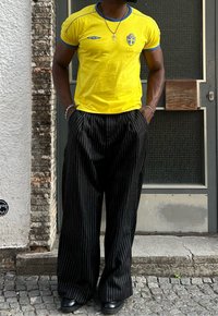 Κίτρινο κοντομάνικο T-shirt με μπλε λεπτομέρειες και σουηδικό έμβλημα, σε συνδυασμό με μαύρο ριγέ φαρδύ παντελόνι και μαύρες μπότες.