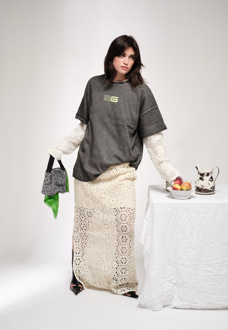 Jeune femme vêtue de vêtements superposés tient un petit sac à main et s’appuie sur une table avec un bol de pommes et une cruche en céramique.