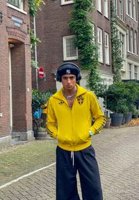 Κίτρινο μπουφάν με φερμουάρ και μπλε λεπτομέρειες και έμβλημα, μαύρο φαρδύ παντελόνι, ακουστικά και μαύρη σκούφια. Αστική τοποθεσία με πράσινα φυτά.