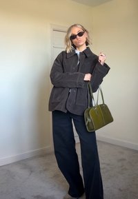 Jachetă din suede maro peste un top alb, asociată cu pantaloni negri lărgi. Ține o geantă verde cu două mânere. Ochelarii de soare completează ținuta.