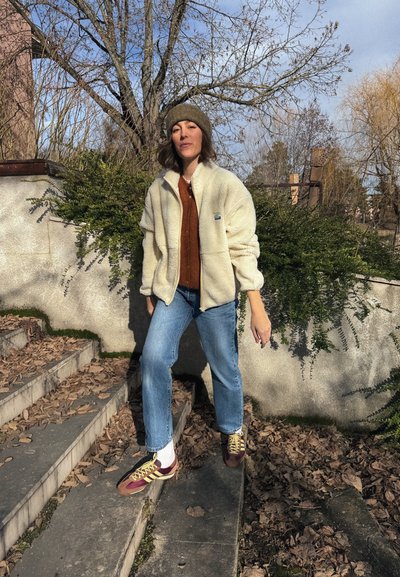 Mujer con chaqueta de forro polar crema, gorro verde, suéter naranja, jeans y zapatillas, de pie en unos escalones al aire libre cubiertos de hojas, con árboles desnudos al fondo.