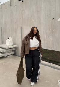 Brązowa skórzana kurtka, szara krótka bluza z kapturem, czarne spodnie dresowe z białymi paskami oraz teksturowana ciemnozielona torba. Nowoczesne wnętrze.