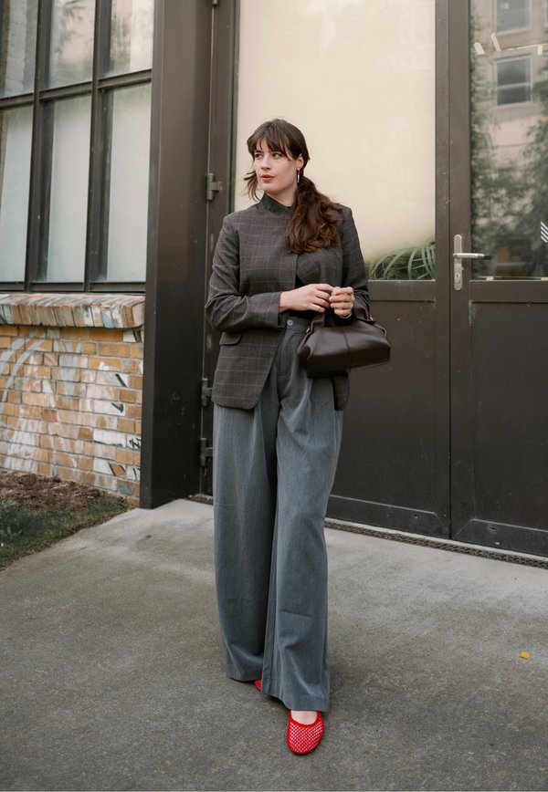 Mujer con blazer a cuadros gris y pantalones anchos grises sosteniendo un bolso marrón, de pie al aire libre junto a una puerta de vidrio, con zapatos rojos.