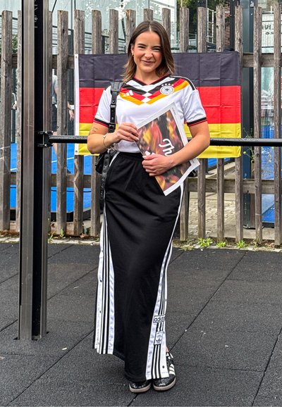 Joven mujer que lleva una camiseta de fútbol alemana y una falda negra de Adidas, está sonriendo, sosteniendo una revista, con una bandera alemana detrás de ella.