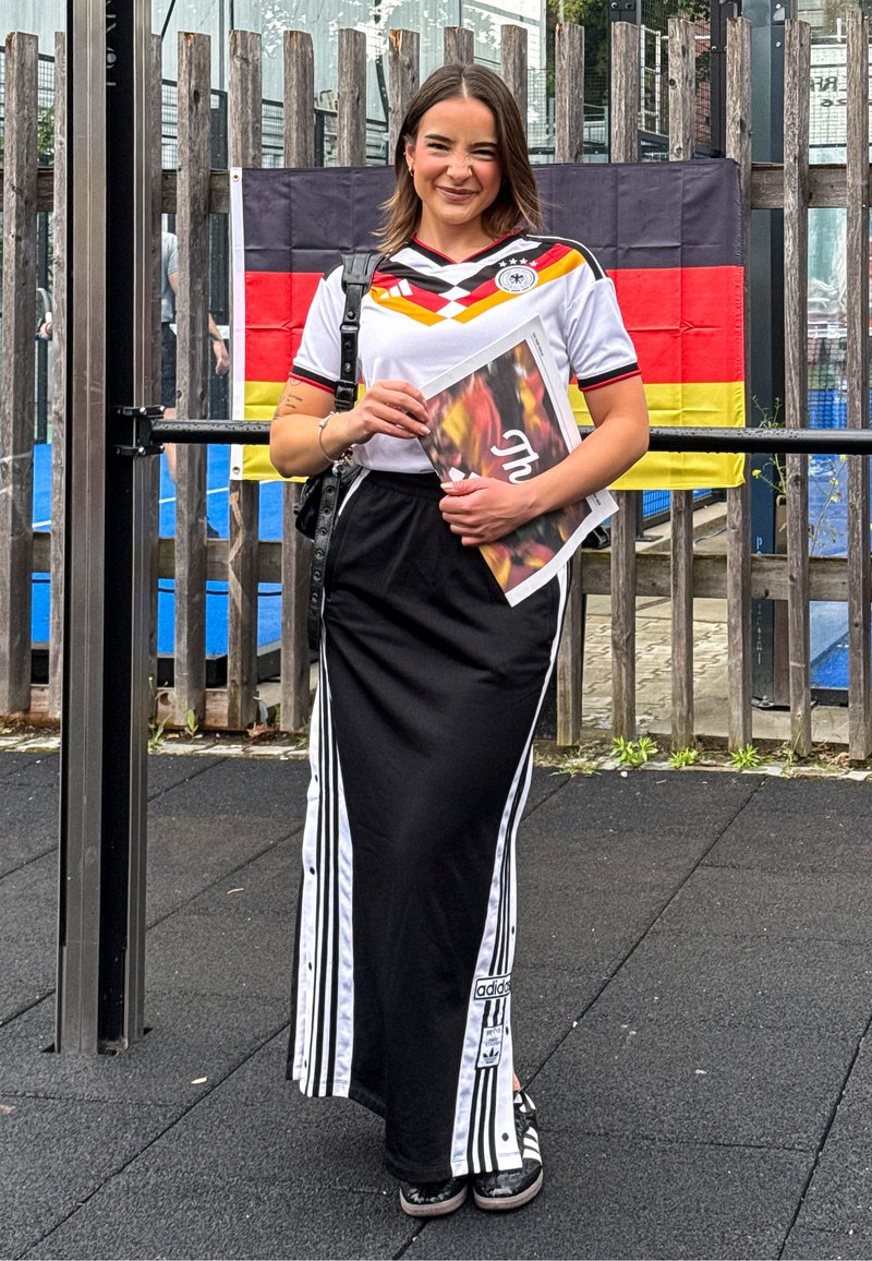 Jeune femme portant un maillot de football allemand et une jupe noire Adidas, souriante, tenant un magazine, avec un drapeau allemand derrière elle.