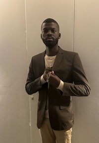 Homme à la barbe taillée et aux boucles d'oreilles ajuste un blazer sombre sur une chemise claire, debout contre un mur beige uni.