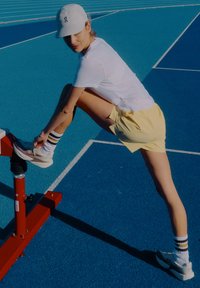 Sporta apavi gaiši pelēkā krāsā ar baltu zolī, kopā ar dzelteniem šortiem un baltu T-kreklu. Cepure ar logotipu. Fons: zils skrejceļa segums.