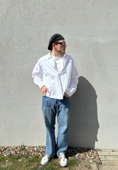 Camisa blanca de gran tamaño con botones sobre una camiseta gris claro, combinada con pantalones vaqueros sueltos azules y zapatillas blancas. Lleva un boina negra y gafas de sol.
