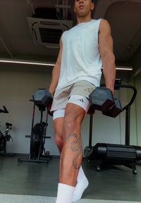 Haltères dans les mains, portant un haut blanc sans manches et un short beige à motif. Tatouage notable sur la jambe, debout sur le sol d'une salle de sport.