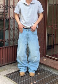 Jeans de ganga azul-claro com um corte à solta, apresentando grandes bolsos e um design de perna reta, combinados com uma camisola polo cinza e botas castanhas.