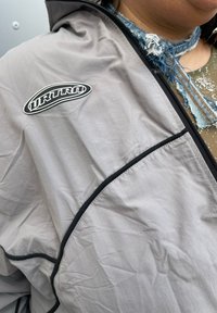 Šedá lehká bunda se zipem, černým lemováním a výraznou černou nášivkou s logem. Materiál vypadá mírně pomačkaně a má hladký povrch.