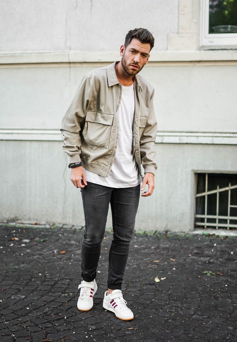 Beige lightweight jacket with two front pockets over a white t-shirt, paired with dark slim-fit jeans and white sneakers with red stripes.