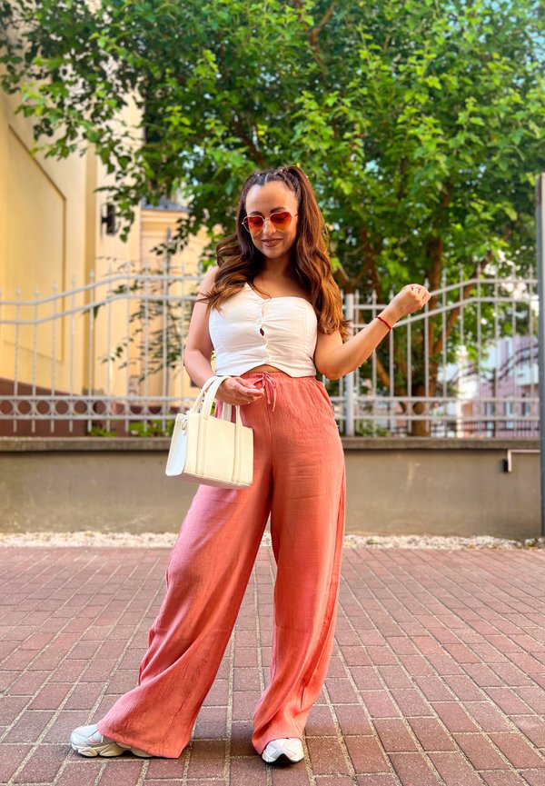 Model wearing a white strapless top and coral wide-leg trousers. She holds a small white handbag, accessorised with round sunglasses and white trainers.