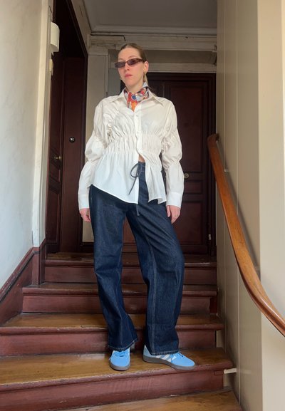 Blusa blanca recogida con mangas largas, pantalones de mezclilla anchos de color oscuro, zapatos azul claro y una bufanda de colores con estampado alrededor del cuello.