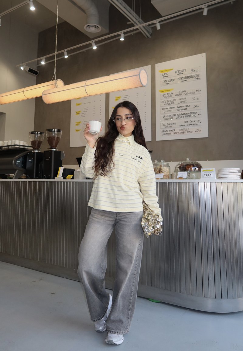 Jeune femme en tenue décontractée tenant une tasse de café à l'intérieur d'un café moderne avec un comptoir en métal et des panneaux de menu illuminés.