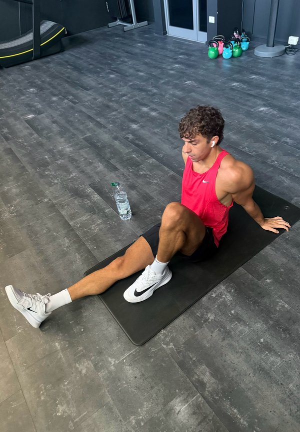 Hombre con camiseta sin mangas roja y pantalones cortos negros se estira sobre una esterilla negra en el gimnasio con pesas rusas y botella de agua cerca.
