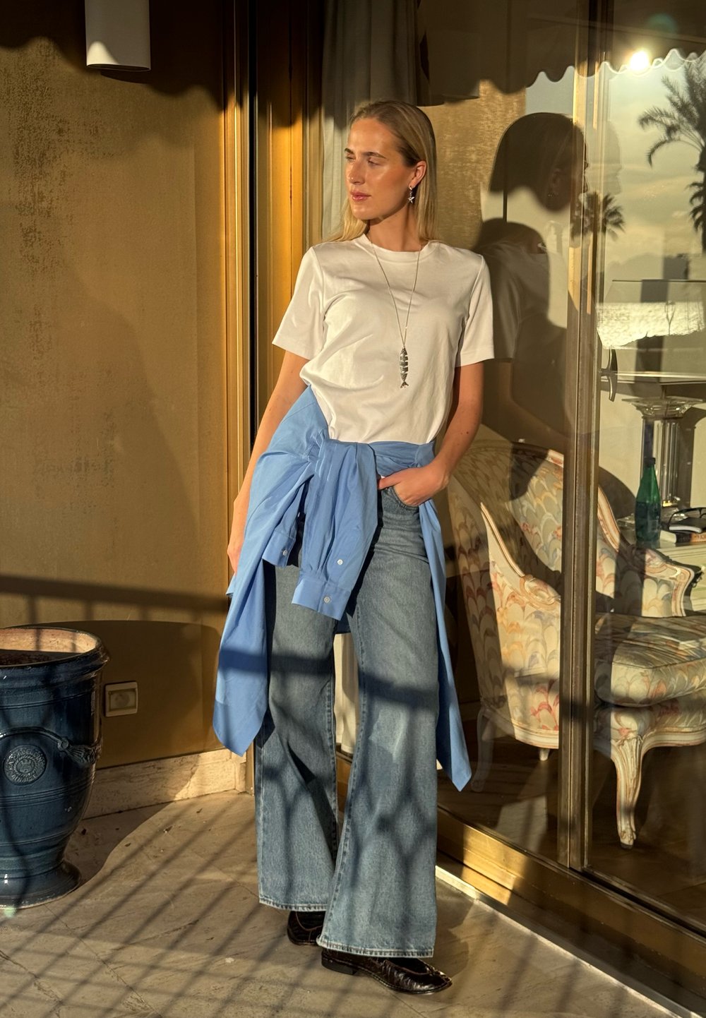 Femme debout sur un balcon ensoleillé, portant un t-shirt blanc, un jean pattes d'éléphant, une chemise bleue nouée autour de la taille et un collier avec un pendentif en forme de poisson.