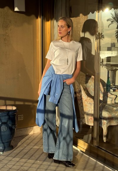 Mujer de pie en un balcón iluminado por el sol, vistiendo una camiseta blanca, jeans de pierna ancha, camisa azul atada a la cintura y un collar con colgante de pez.