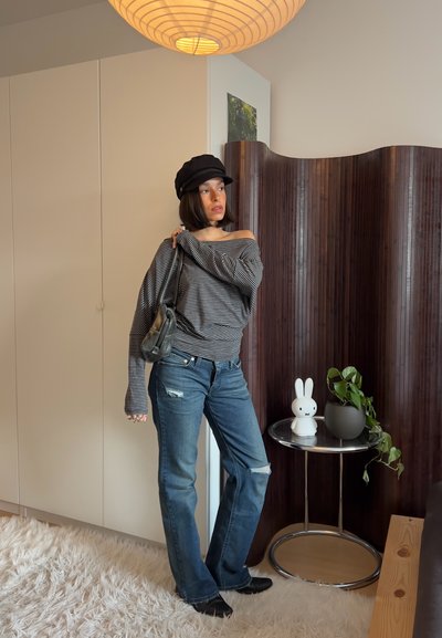 Mujer con suéter a rayas con hombros descubiertos, vaqueros rotos y gorra negra, de pie cerca de una mesa con una lámpara de conejito blanca y una planta en maceta en una habitación moderna.