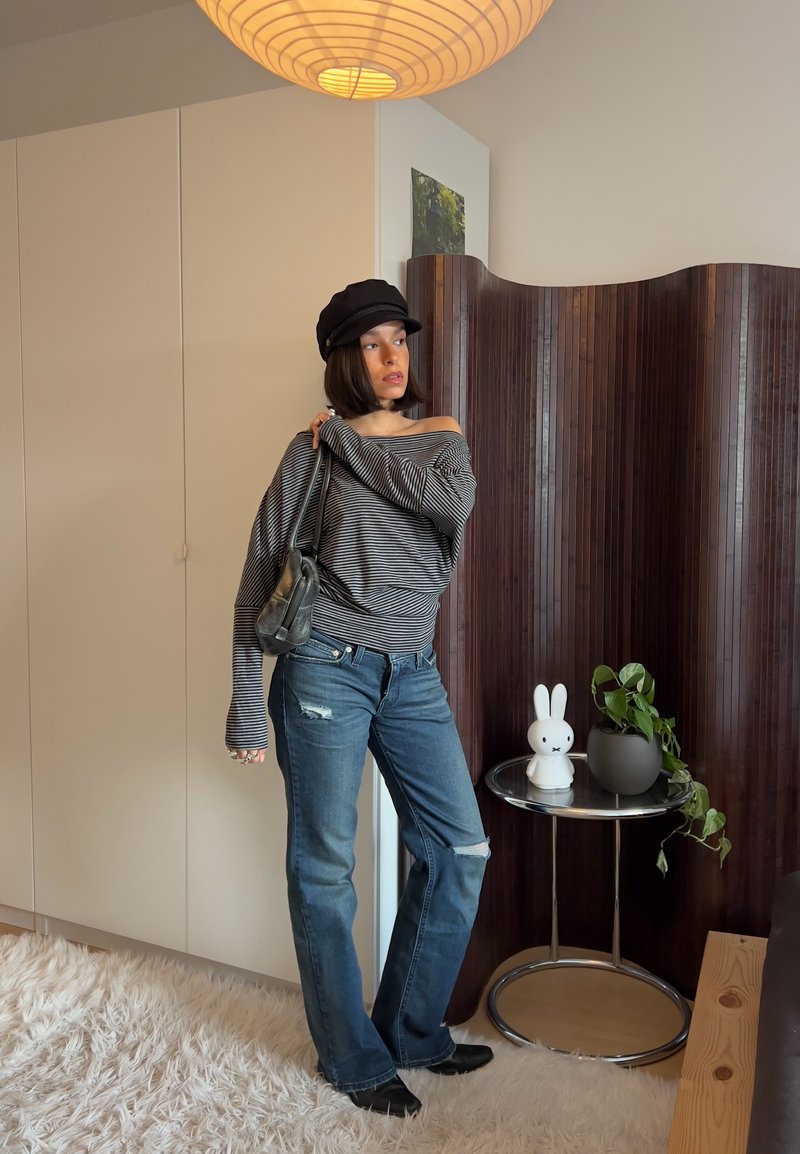 Donna con maglione a righe con spalle scoperte, jeans strappati e cappellino nero in piedi vicino a un tavolo con una lampada a coniglietto bianca e una pianta in vaso in una stanza moderna.