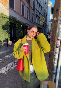 Manteau vert surdimensionné texturé, haut marron boutonné, jupe noire, sac rouge, tenant une tasse rouge, avec des lunettes de soleil et des boucles d'oreilles créoles. Fond de rue pavée.