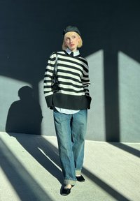 Μαύρο και άσπρο ριγέ φαρδύ πουλόβερ, άσπρη μπλούζα από κάτω, χαλαρά μπλε τζιν, μαύρο μπερές και μαύρα παπούτσια σε λείο τσιμεντένιο έδαφος.