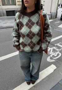 Maglione argyle verde e marrone, lavorato a maglia con una finitura testurizzata, abbinato a jeans a gamba larga azzurri e mocassini marroni. Borsa in pelle marrone.