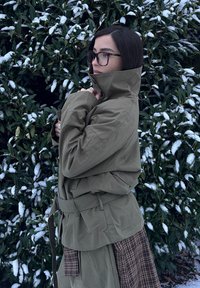 Veste oversize vert olive avec col montant et ceinture, portée sur une jupe marron à motifs, au milieu d'un feuillage vert et d'accents de neige.