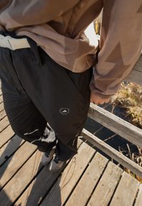 Pessoa com um fleece bege e calças pretas com logótipo está em uma ponte de madeira, segurando o corrimão, ao lado de água com plantas secas.