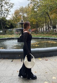Robe en tricot noire à manches longues, avec un ourlet évasé et des épaules dénudées. Tient un sac à main blanc en forme de croissant. Chaussures à plateforme noires. Fond de fontaine.