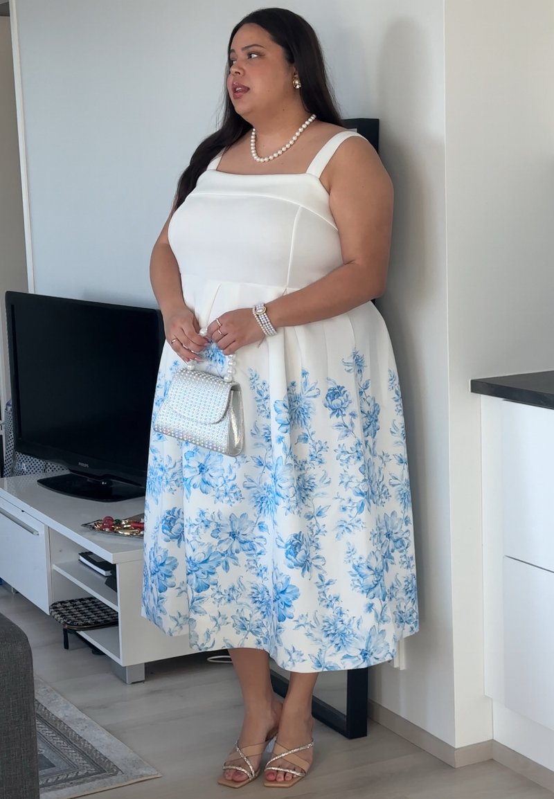 Femme debout à l'intérieur portant une robe blanche avec imprimé floral bleu, un collier de perles, un bracelet, tenant un petit sac à main et portant des sandales.