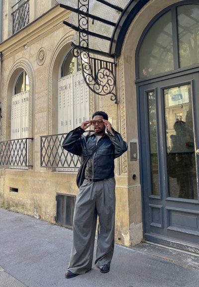 Chaqueta de cuero negra, camisa blanca, pantalones de pierna ancha grises y gafas de sol elegantes. De pie junto a un edificio vintage con elementos decorativos.