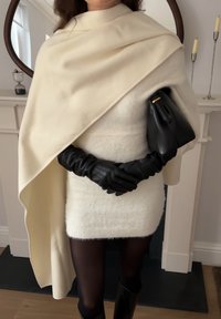 Femme portant une robe en maille blanche et un châle assorti, des gants en cuir noir, des collants noirs, tenant un sac à main noir devant une cheminée blanche.