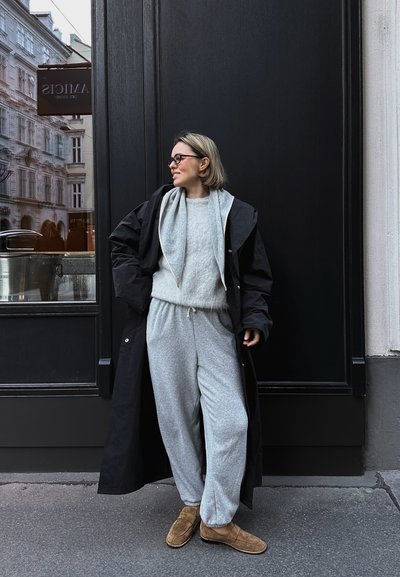 Abrigo negro largo sobre un suéter gris claro y pantalones de chándal a juego. Zapatos de ante marrón tipo slip-on. Gafas de montura negra. Fondo oscuro.