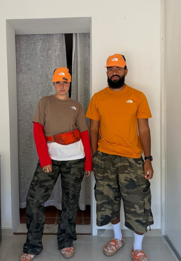 Dos personas están de pie en el interior llevando gorras naranjas de North Face, pantalones de camuflaje, zapatos naranjas y camisetas informales con expresiones neutrales.