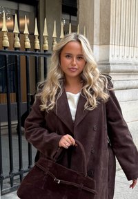 Brun semsket dobbelspent kåpe, hvit topp under, holder en mørk brun semsket håndveske med strukturert design og metallhardware-detaljer.