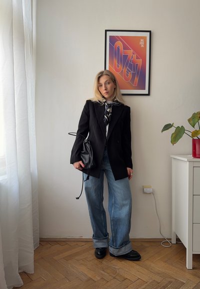 Mujer con cabello rubio que lleva una chaqueta negra, bufanda, vaqueros anchos y zapatos negros, de pie en una habitación con un póster enmarcado y una maceta.