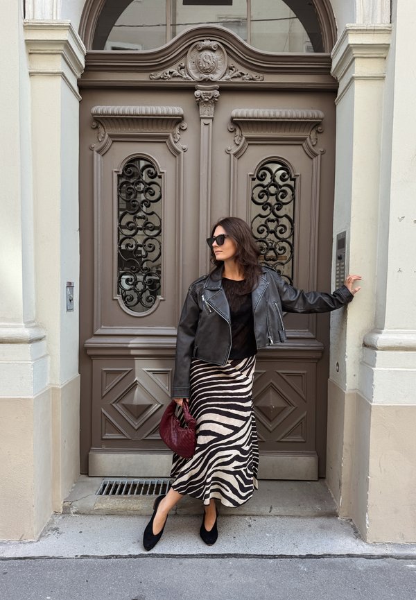 Mujer con chaqueta de cuero negra y falda con rayas de cebra, de pie junto a una puerta marrón ornamentada, sosteniendo un bolso rojo y mirando de lado.