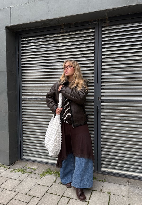 Ung kvinde med briller iført mørk jakke, stribet top, lagdelt nederdel og brede jeans, der holder en stor hvid tekstureret taske ved en metalrulleport.